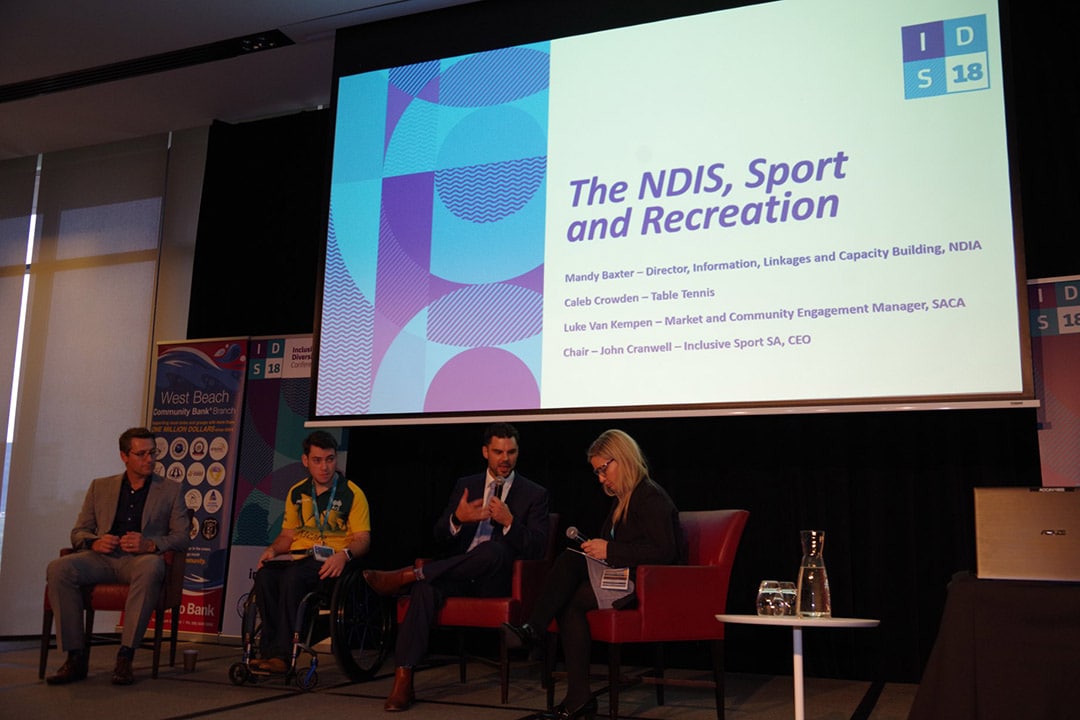 a group of four adults sit on a stage in front of a screen that reads The NDIS Sport and Recreation