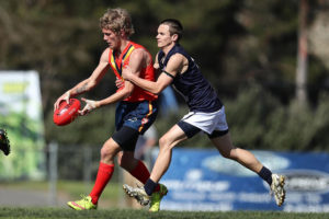 A South Australian football player is holding the football mid game, a Victorian player has begun to tackle him.