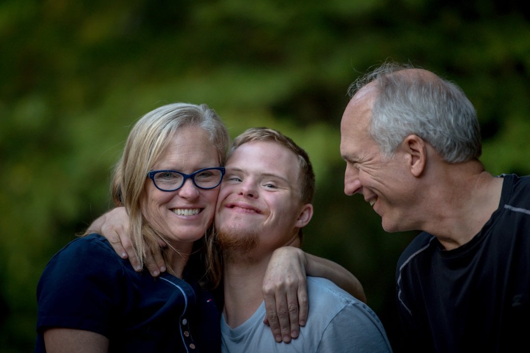 A family of three a couple and their young adult son stand outside. The son is hugging his mother whilst the father smiles at the two of them.
