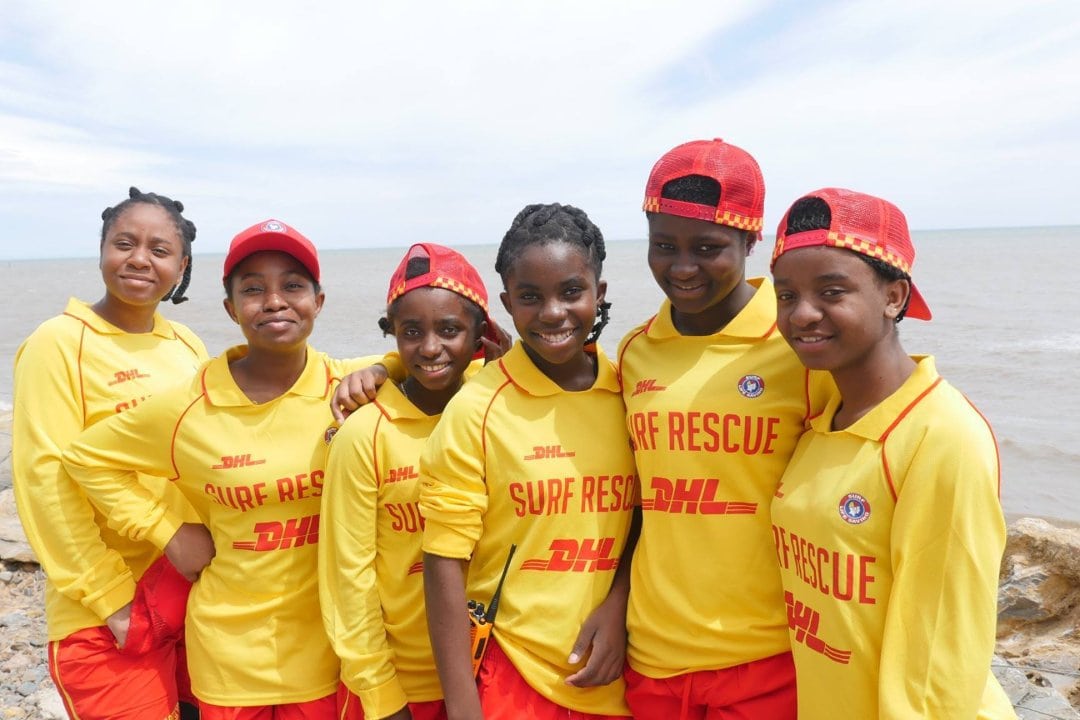 a group of six teenage and young women stand together on the beach wearing lifesaving uniforms