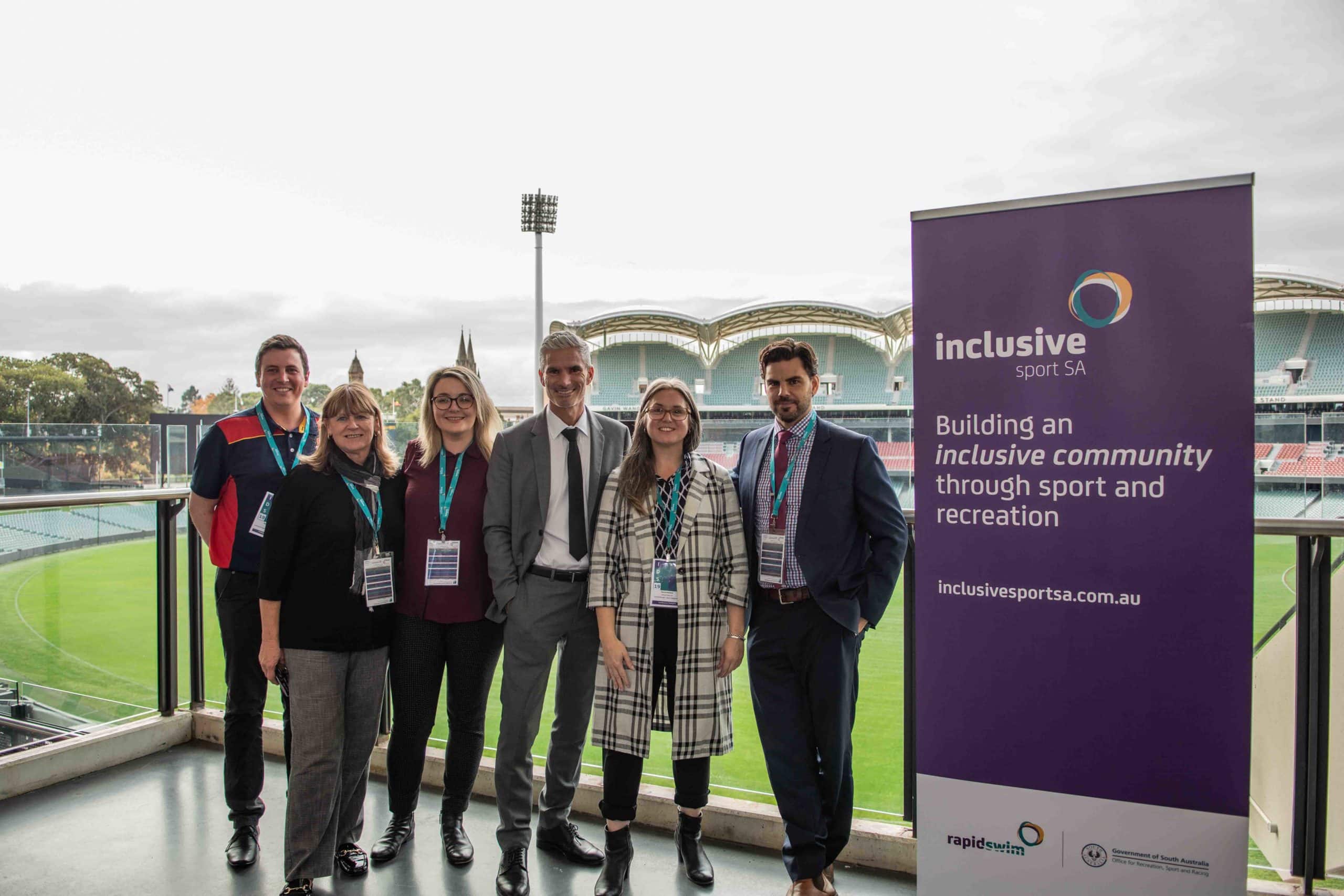 Group of six Active inclusion staff and collaborators standing on balcony with Adelaide Oval in the background.