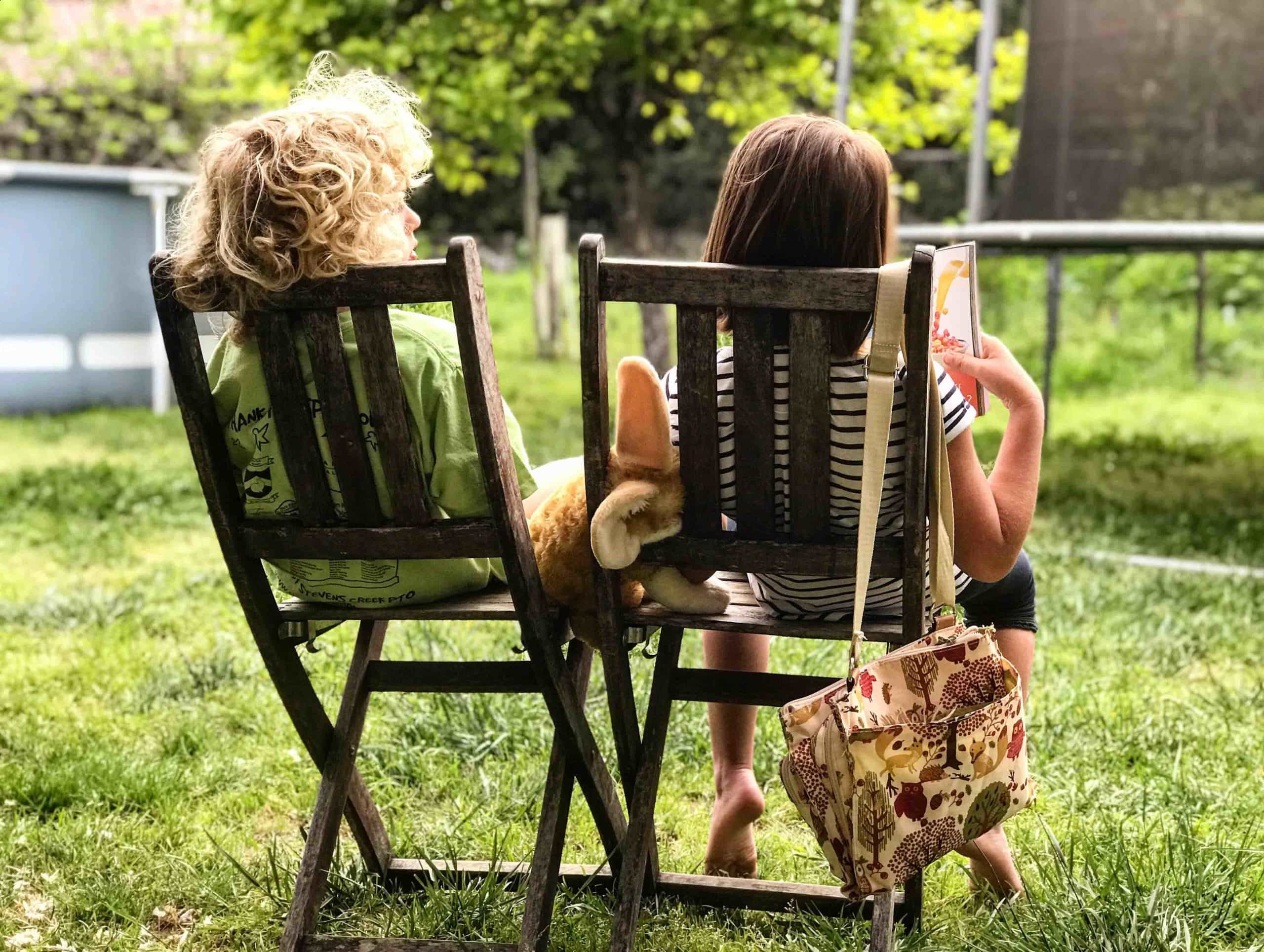 Two children sit on chairs next to each other in the garden. The young boy is talking to the young girl who is reading a book.