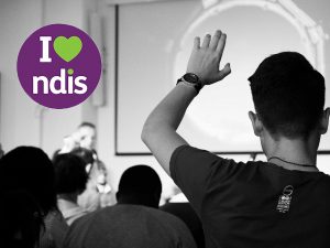 Man at the front of a room of people points to a man who has his hand up to ask a question. The I love NDIS logo appears in the top left corner.