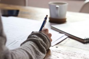 Person wearing a brown jumper is writing on paper at a wooden table in the day time. A coffee cup and notebook are also on the table.