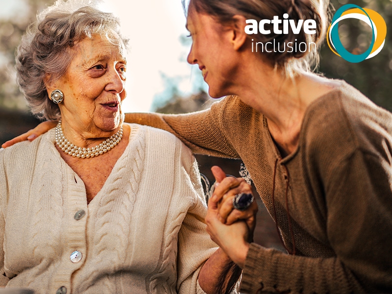 An older woman sits outside smiling. She is talking to a younger woman who has her arm around her and is holding her hand. The Active Inclusion logo is in the top right corner.