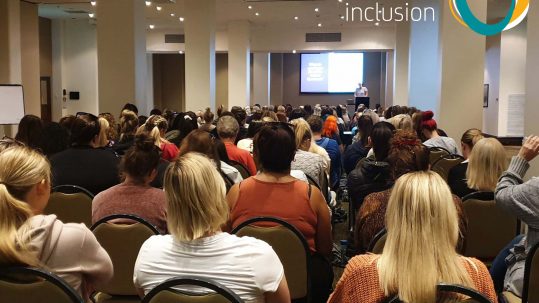 Image shows a room full of people at a conference. At the front of the room is a screen everyone is watching this and the person presenting. The Active Inclusion logo appears in the top right corner of the image.