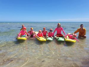 Image of young Seabird Nippers members in the shallow water at the beach on surf life saving boards with their program buddies/instructors.