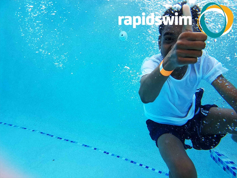 A young boy is underwater smiling and giving a thumbs up gesture. The Rapidswim logo appears in the top right hand corner.